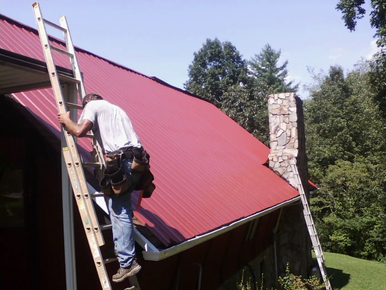 Roofer on a ladder beside a red metal roof and stone chimney — Soffit & Fascia Repair by Roofing Brothers in Usk